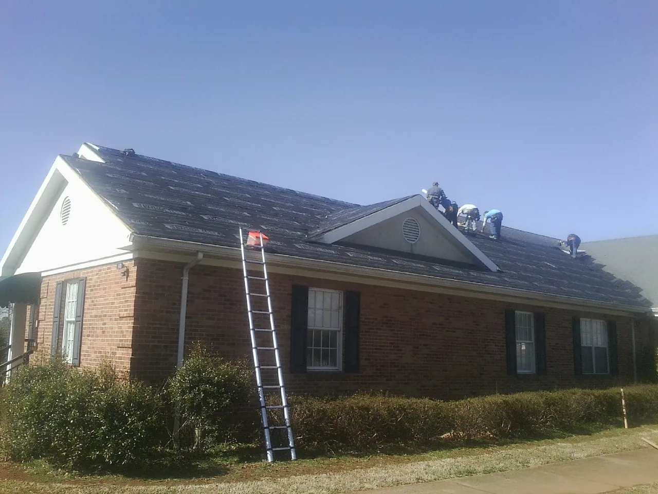 Crew tearing shingles off a brick ranch roof with a ladder set — Emergency Roof Repair work by our crew in Roann