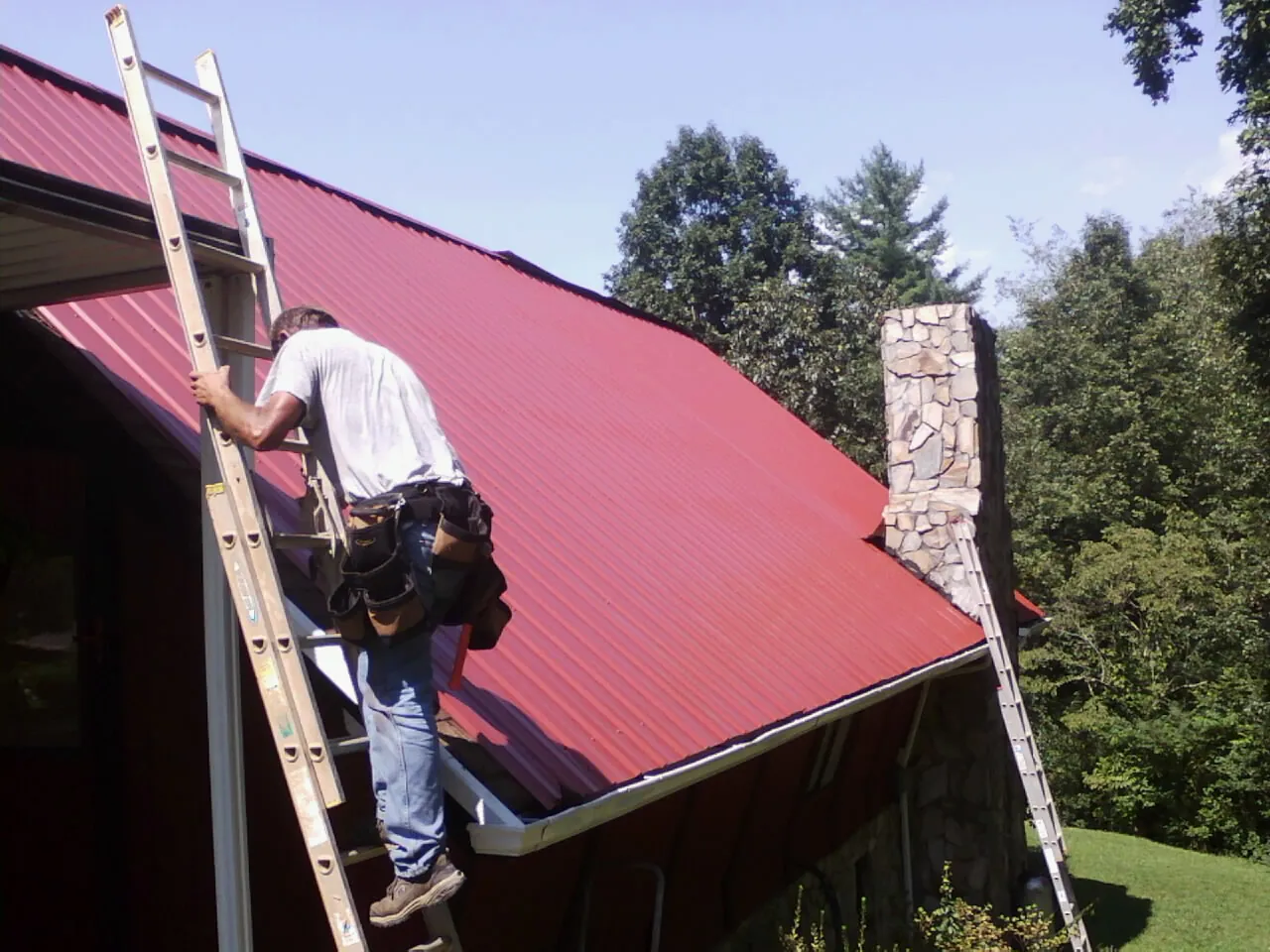 Roofer on a ladder beside a red metal roof and stone chimney — Soffit & Fascia Repair by Roofing Brothers in Roann